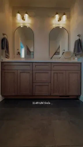 Double-sink bathroom vanity with wood cabinets, arched mirrors, and wall sconces in a warm-toned room