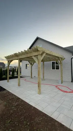 Backyard patio with a wooden pergola beside a white house and paved patio area.