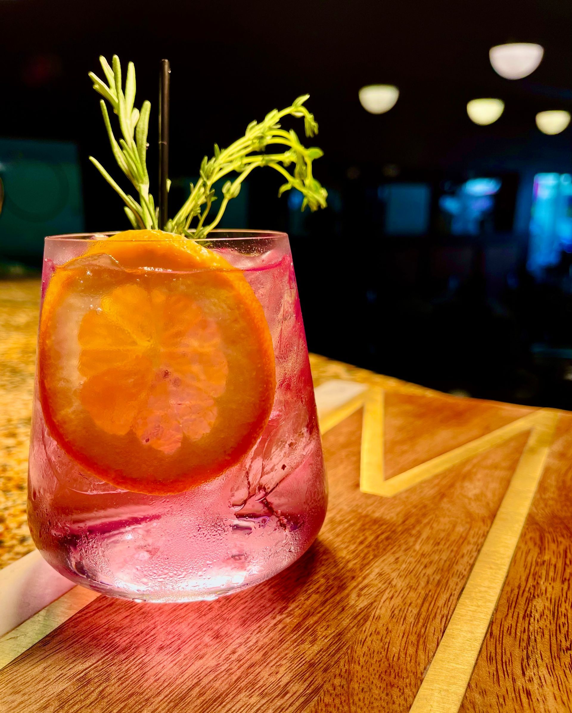 Pink cocktail in a glass, orange slice and rosemary garnish on a wooden bar.