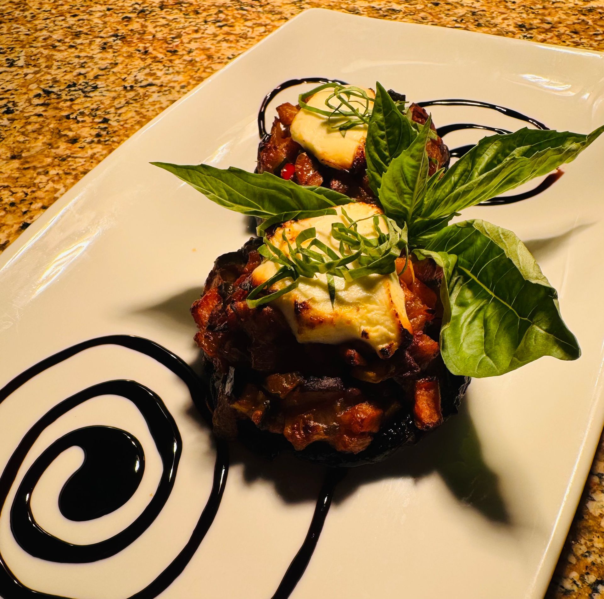 Two stuffed mushrooms topped with goat cheese, basil, and balsamic glaze on a white rectangular plate.