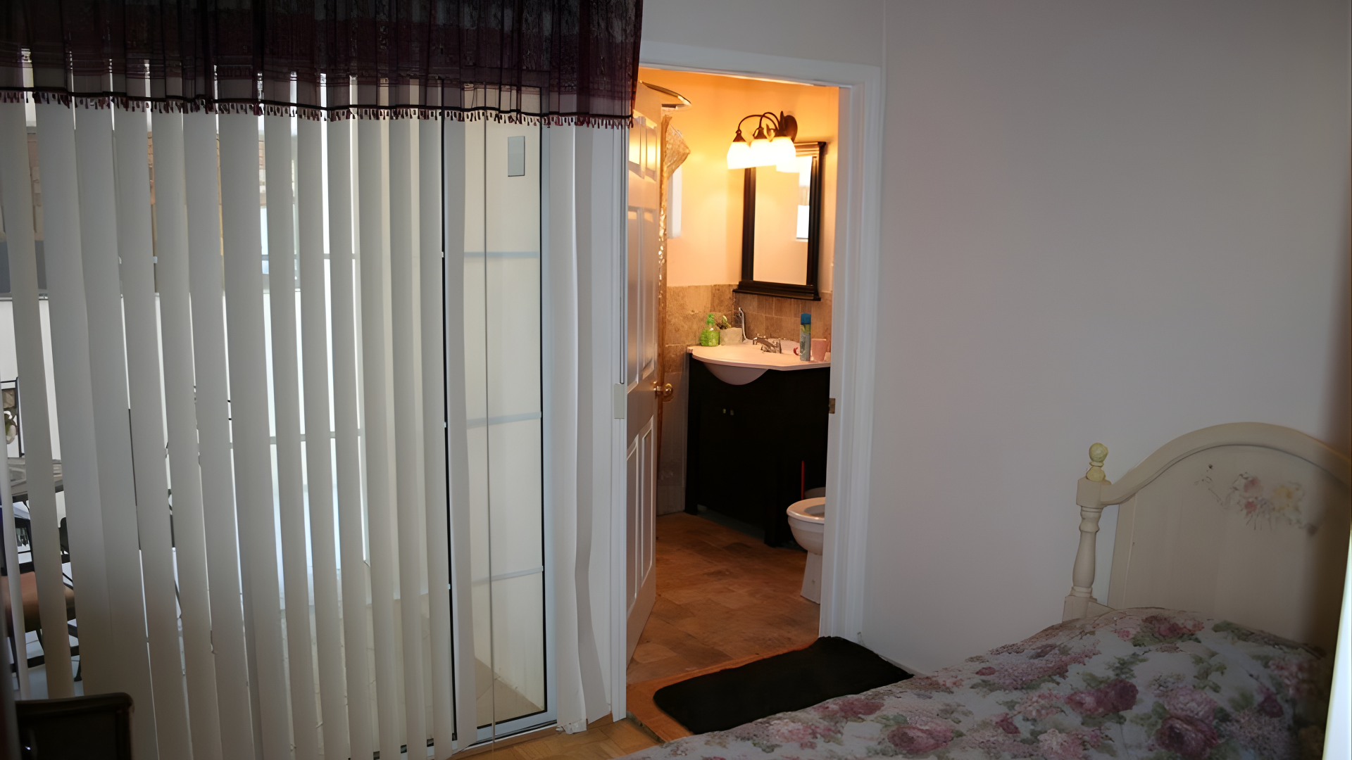 A bedroom with a floral bed, vertical blinds on a glass door, and an open doorway leading to a lit bathroom.
