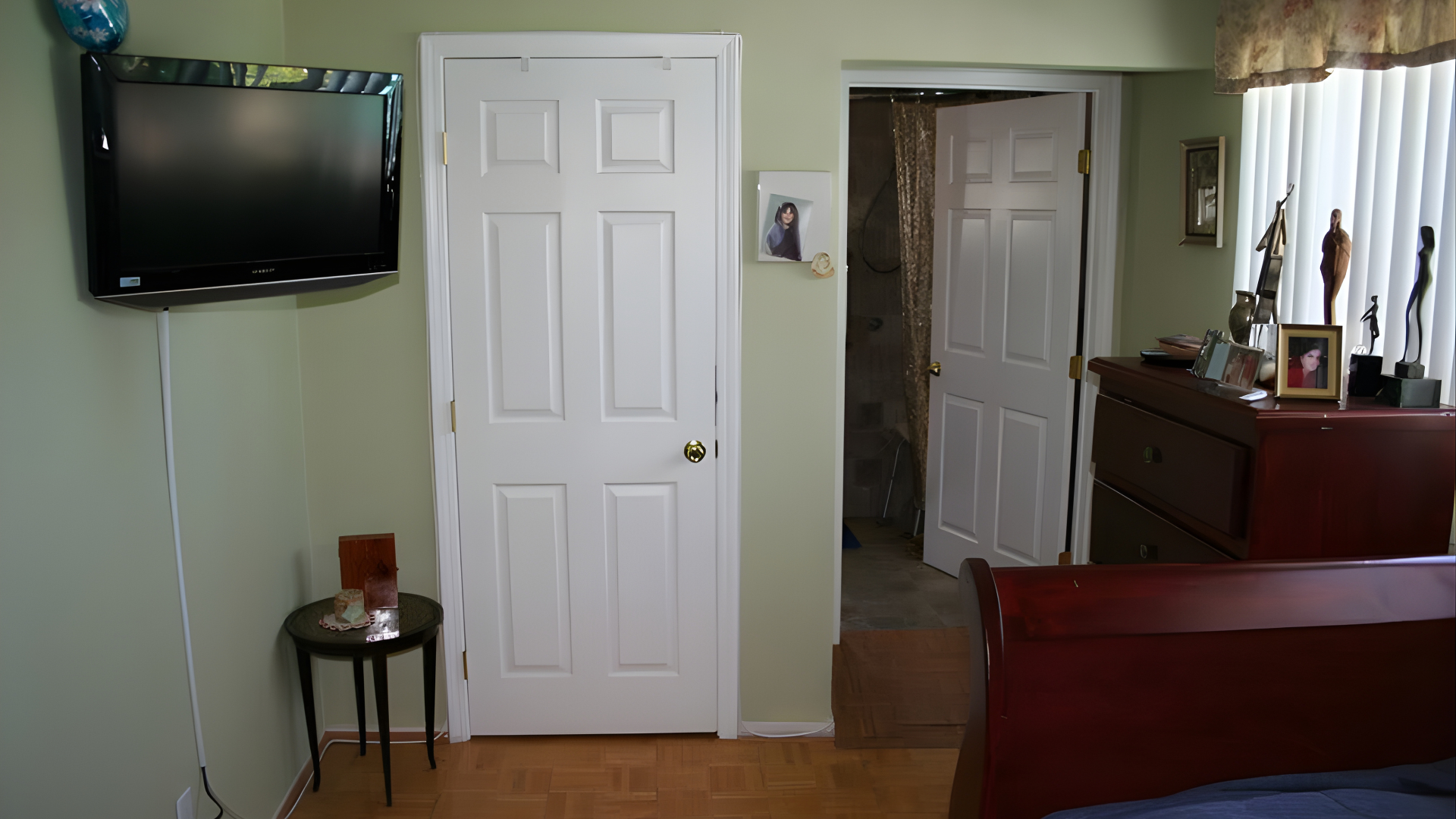 A bedroom corner featuring a wall-mounted TV, a side table, a white door, an open doorway, and a dark wood dresser.