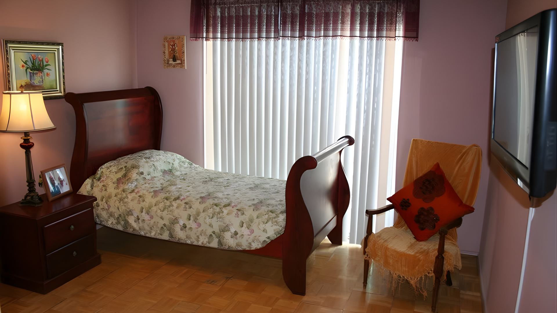 A bedroom with a dark wood bed, patterned bedding, a nightstand with a lamp, a yellow chair, and a wall-mounted TV.