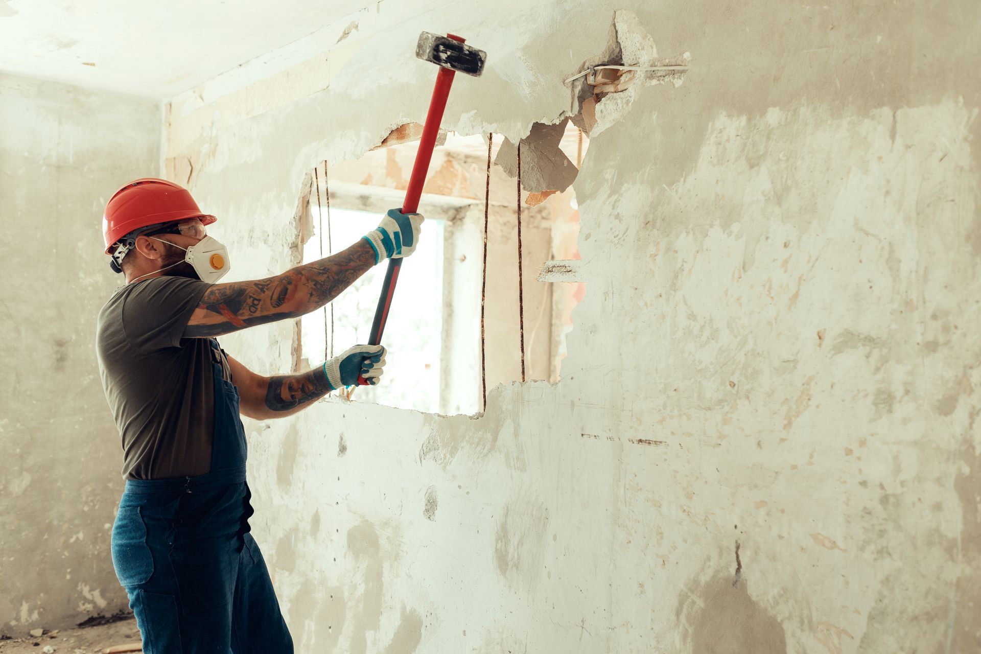 A builder dressed in a protective suit and helmet with a hammer in his hands breaks a cement wall.