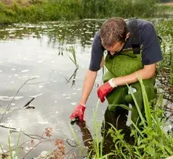 a man in a hard hat and orange vest is taking a sample of water from a pond