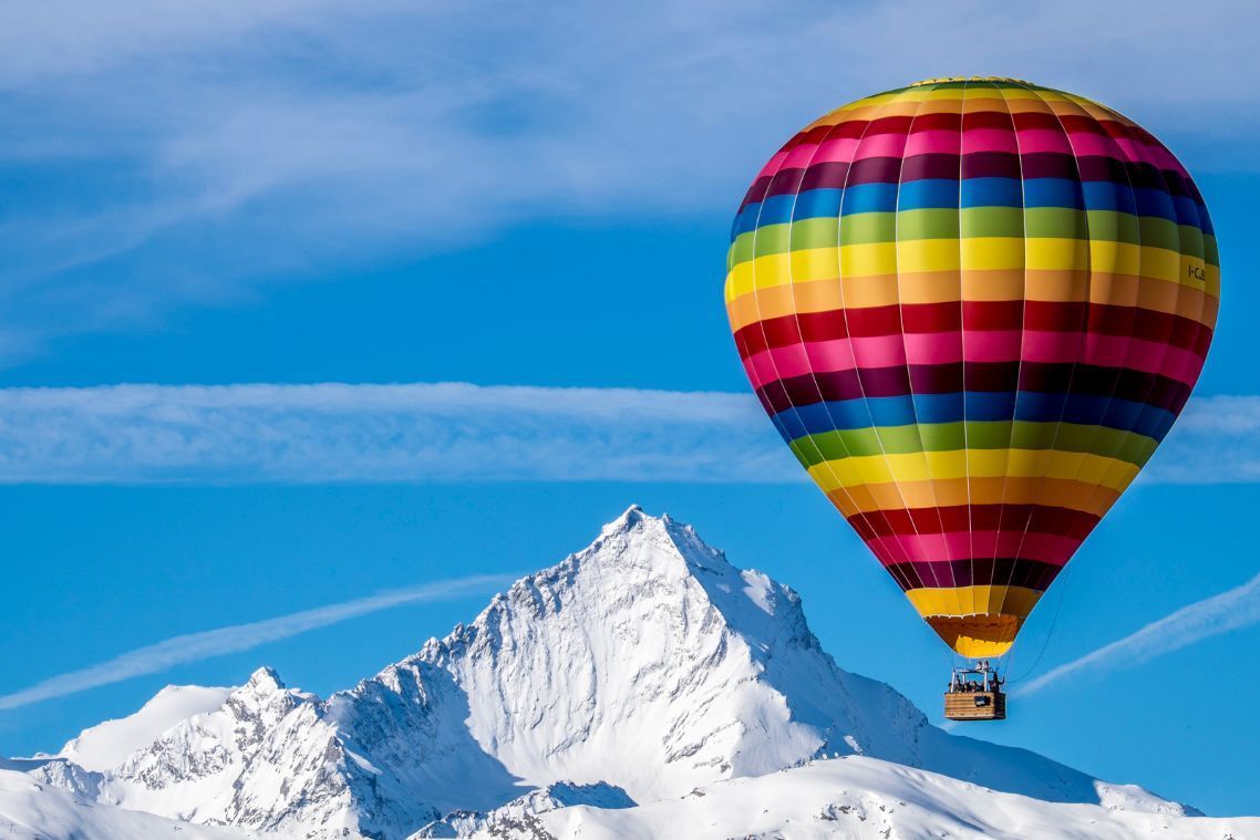 Hot-air balloon flying over the Aosta Valley with snow-covered mountain backdrop.