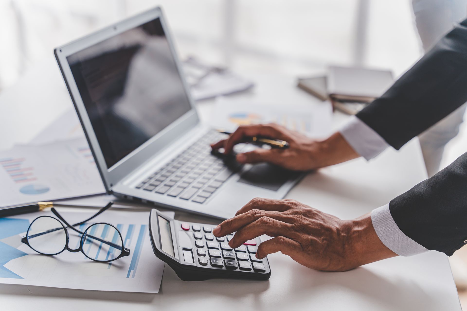 Hands typing on calculator beside laptop and glasses on a white desk