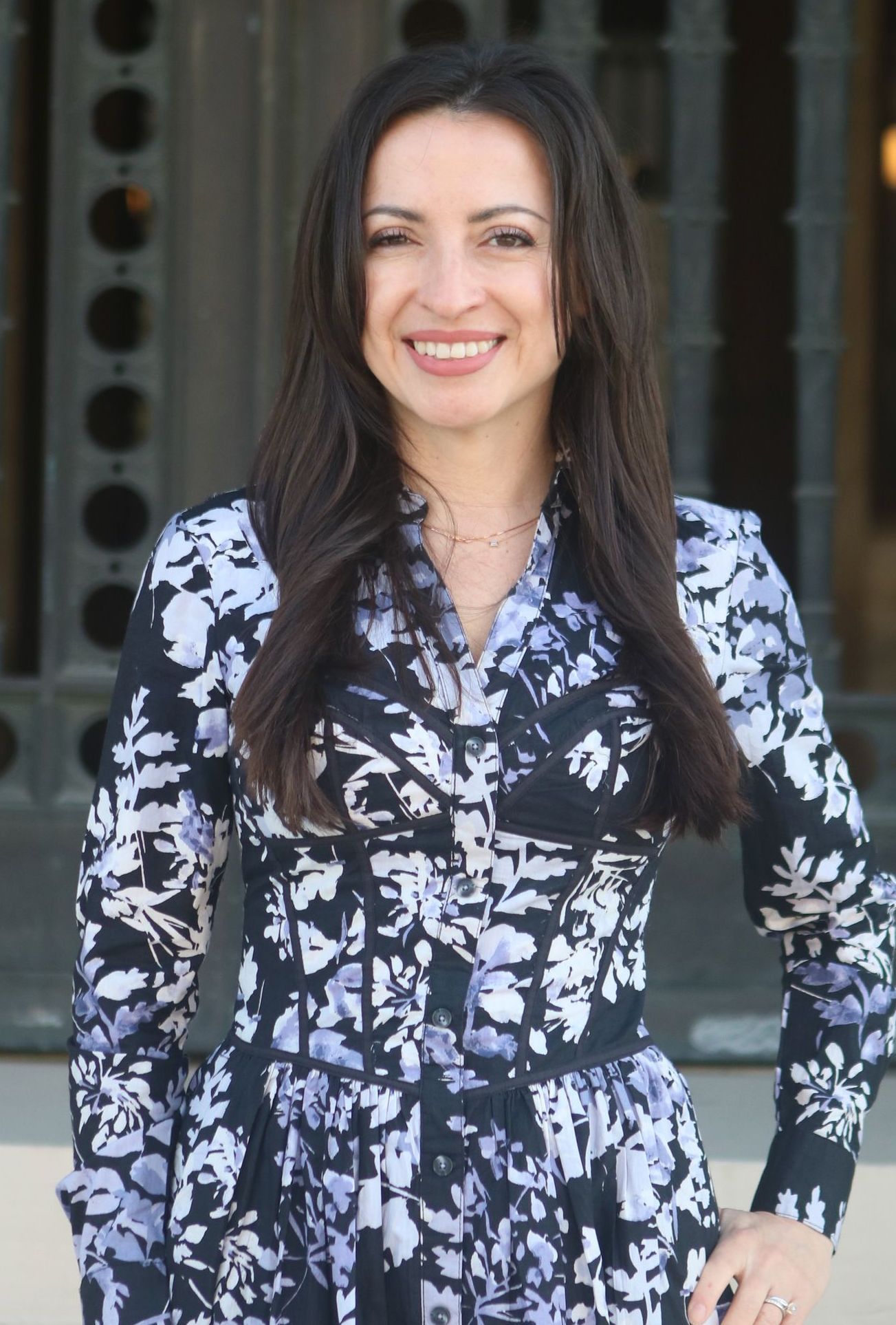 Woman with dark hair smiles in front of a building, wearing a patterned dress.