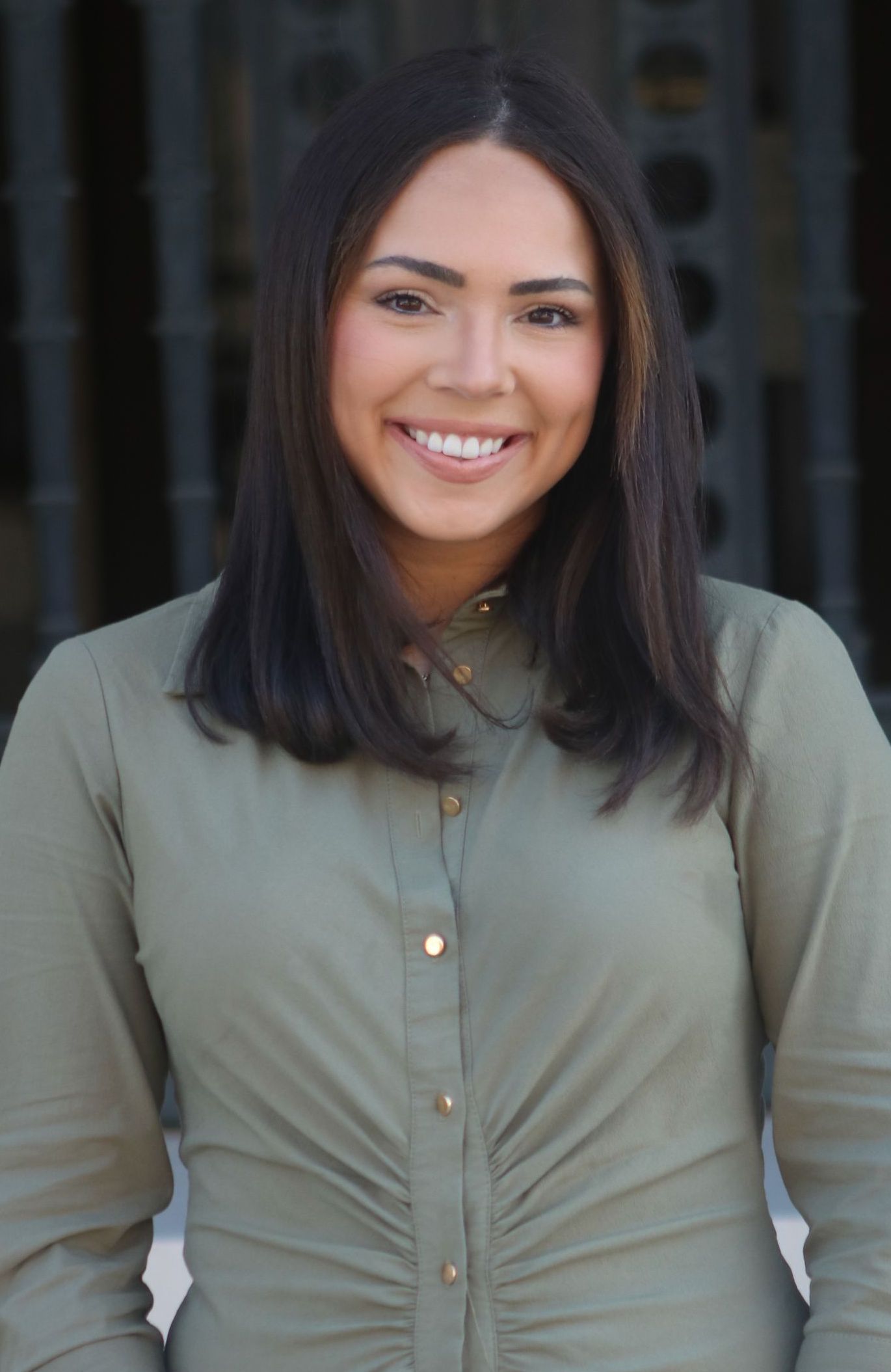 Woman with long brown hair, smiling, wearing a button-down olive green shirt. Outdoors, blurry background.
