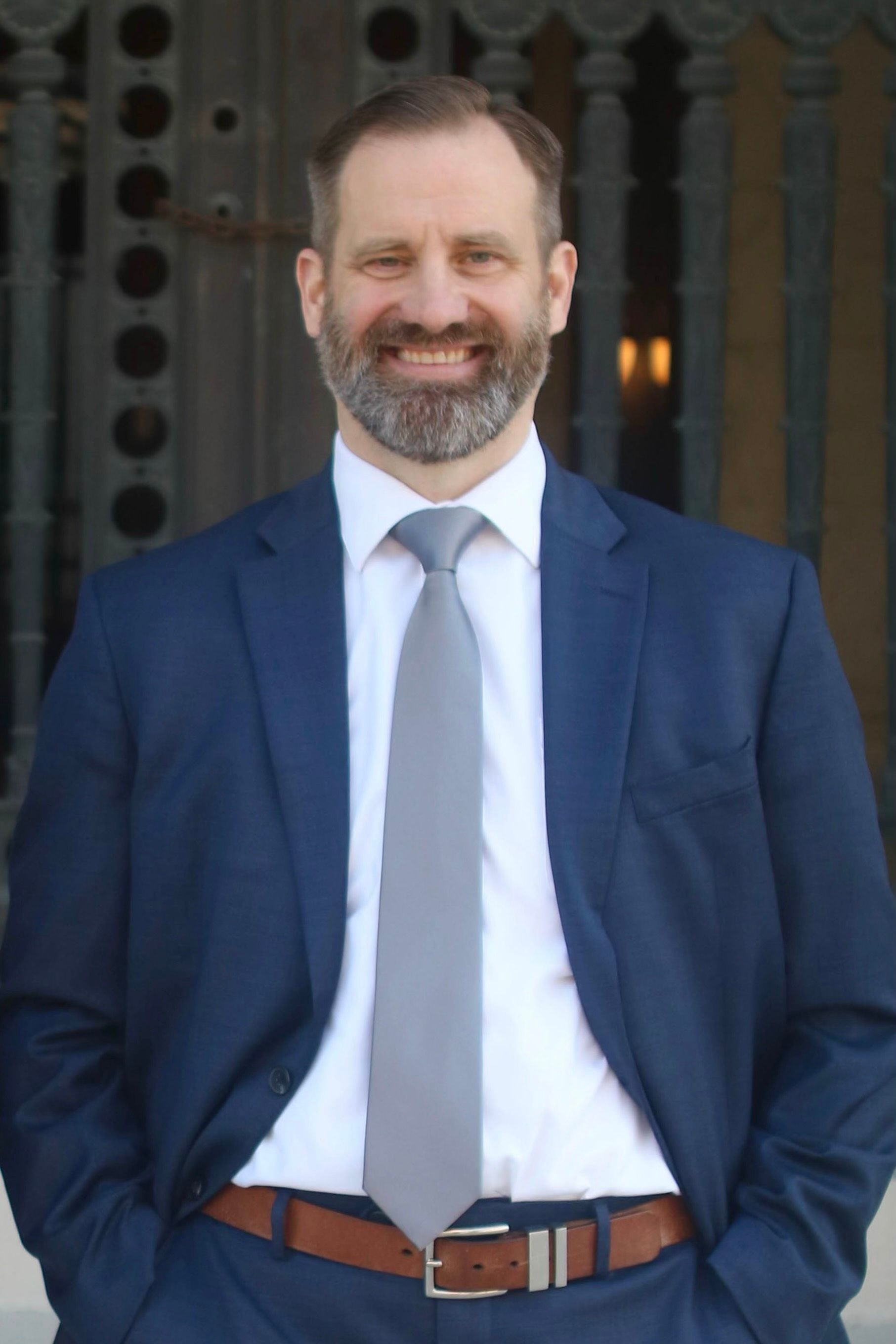 Man in a suit smiling, hands in pockets, standing in front of a building with ornate details.