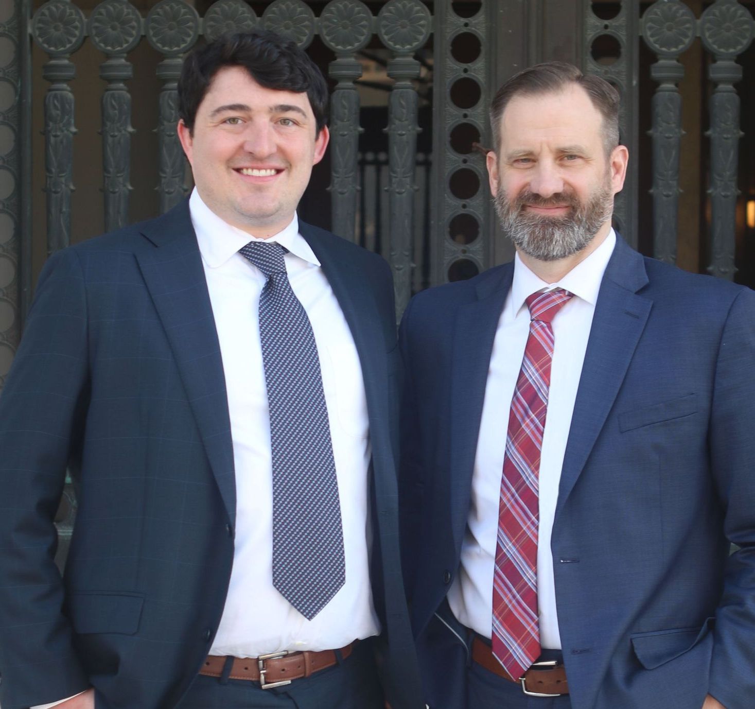 Two men in suits smiling, posing in front of a building's ornate gate. One wears a blue tie, the other a red one.