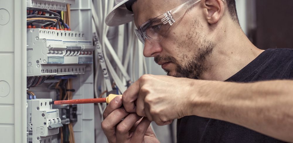 Electricista trabajando en un panel eléctrico con un destornillador, usando gafas de seguridad y una gorra.