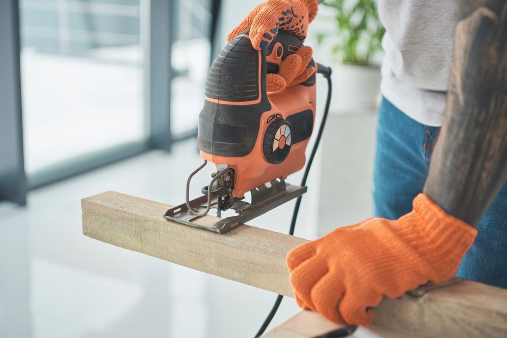 Persona que utiliza una sierra de calar para cortar una tabla de madera, con guantes naranjas.
