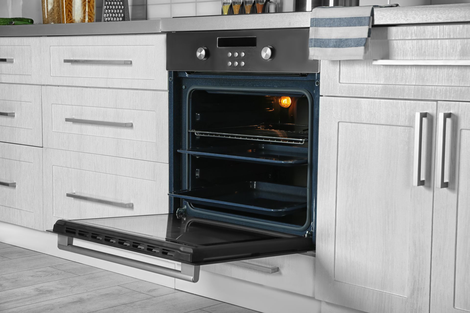 Opened oven built into white cabinets, light on.