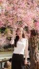 一位身穿白色上衣和黑色裙子的女子站在花園中，粉紅色的花朵簇擁著她。