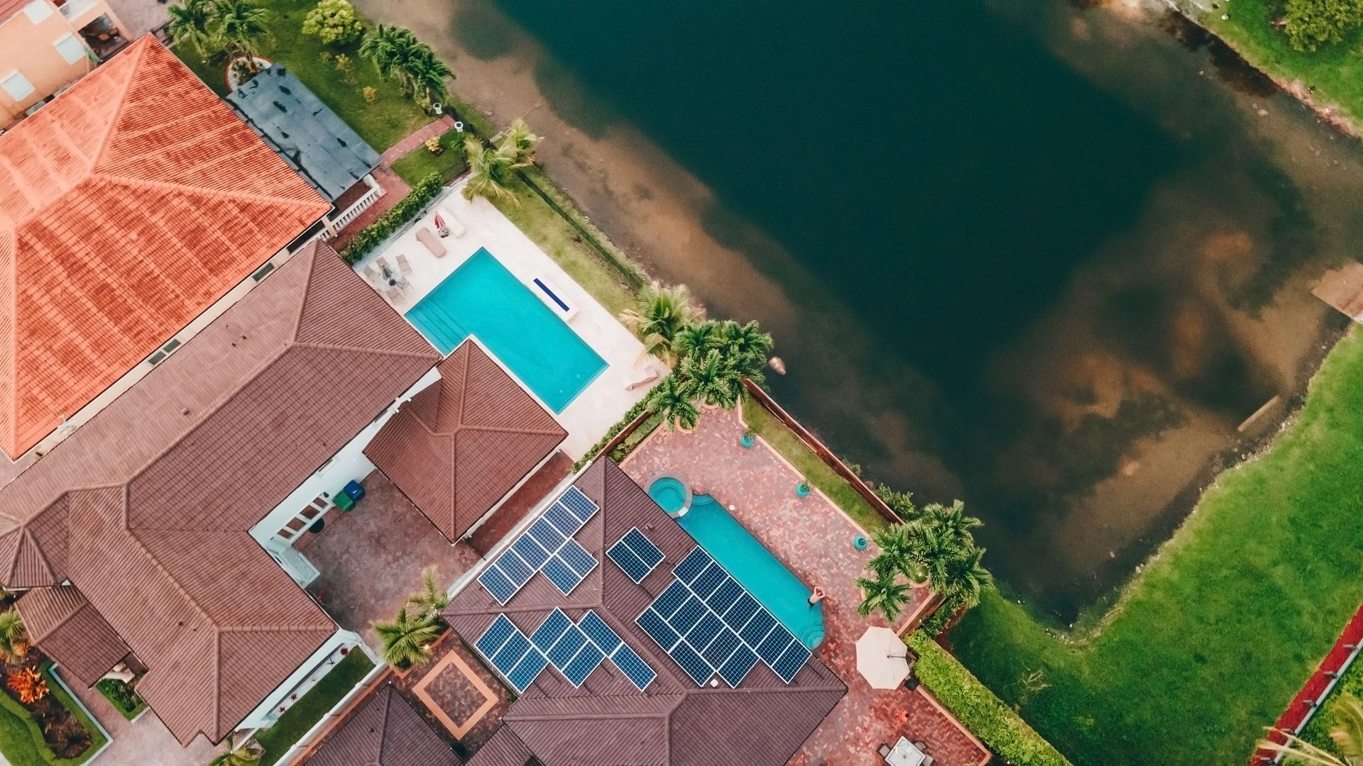 Aerial view of a house with solar panels and a turquoise pool beside a green canal and lawn.