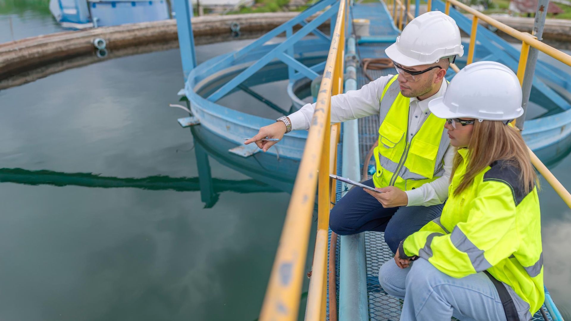 Two workers in hard hats and safety vests inspect water at an industrial facility platform.