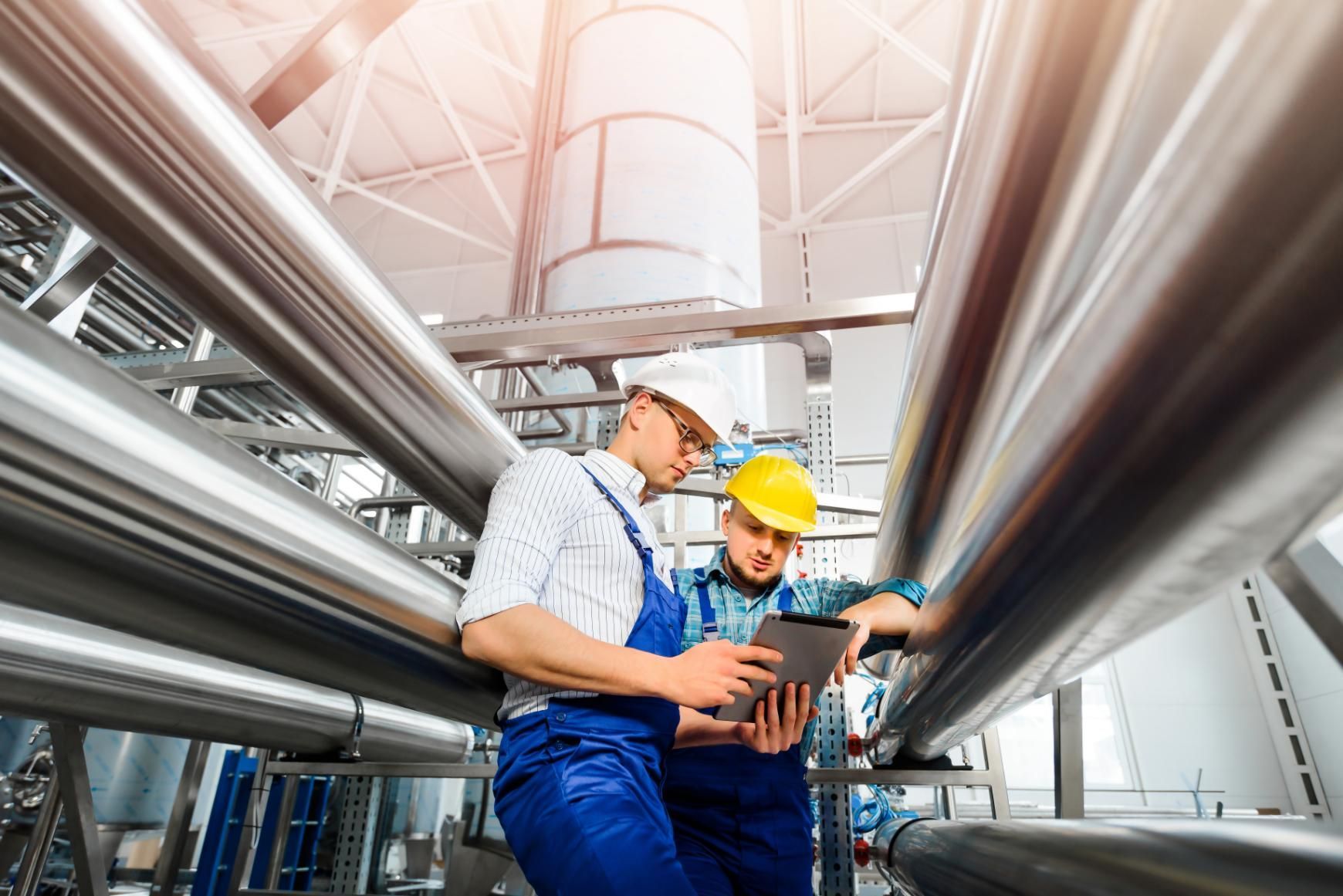 Two workers in hard hats inspect large metal pipes in an industrial facility