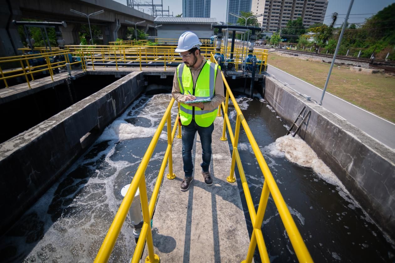 Worker in a hard hat and reflective vest stands on a walkway beside a water treatment channel.