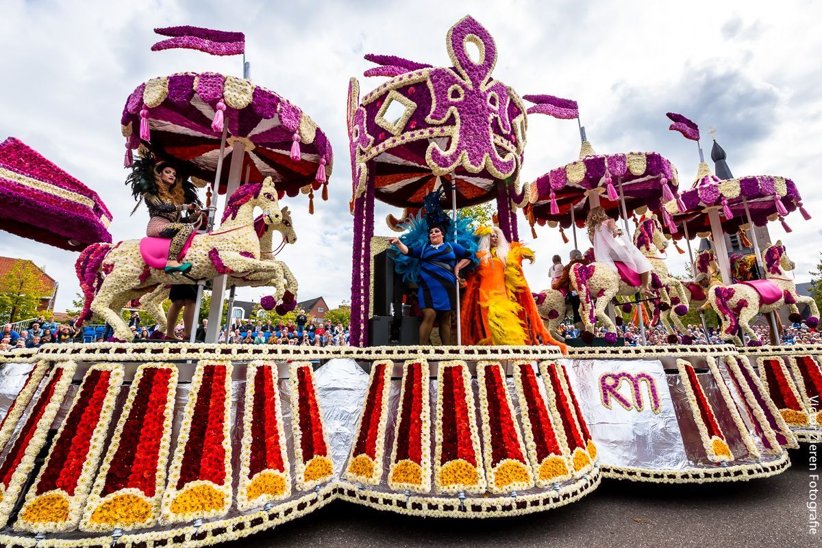 Kleurrijke festivalwagen met paarse versieringen en rood-witte draperieën, waarop artiesten in een parade meelopen.
