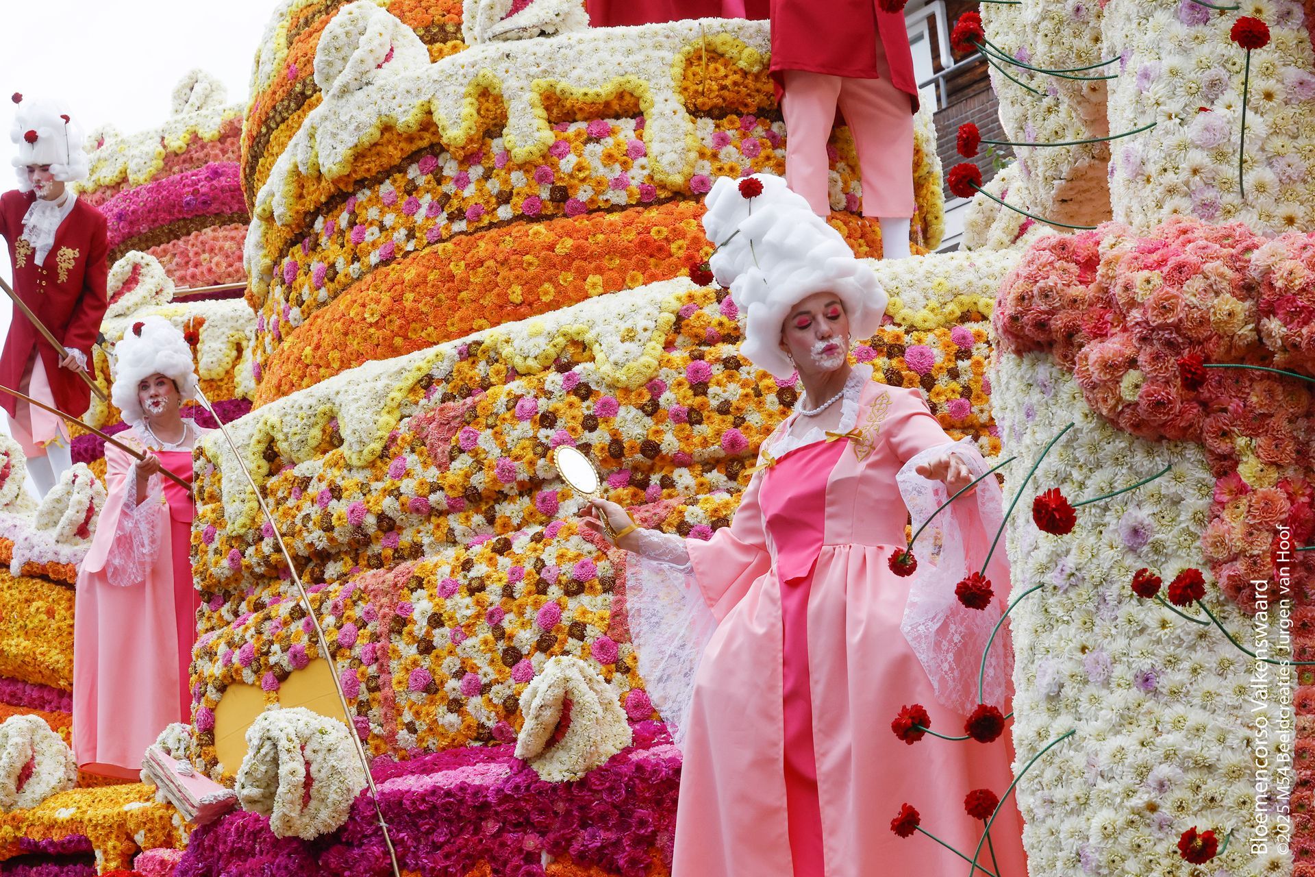 Roze praalwagen met bloemenversiering, artiesten in roze kostuums en witte hoofddoeken te midden van neerdalende bloemen.