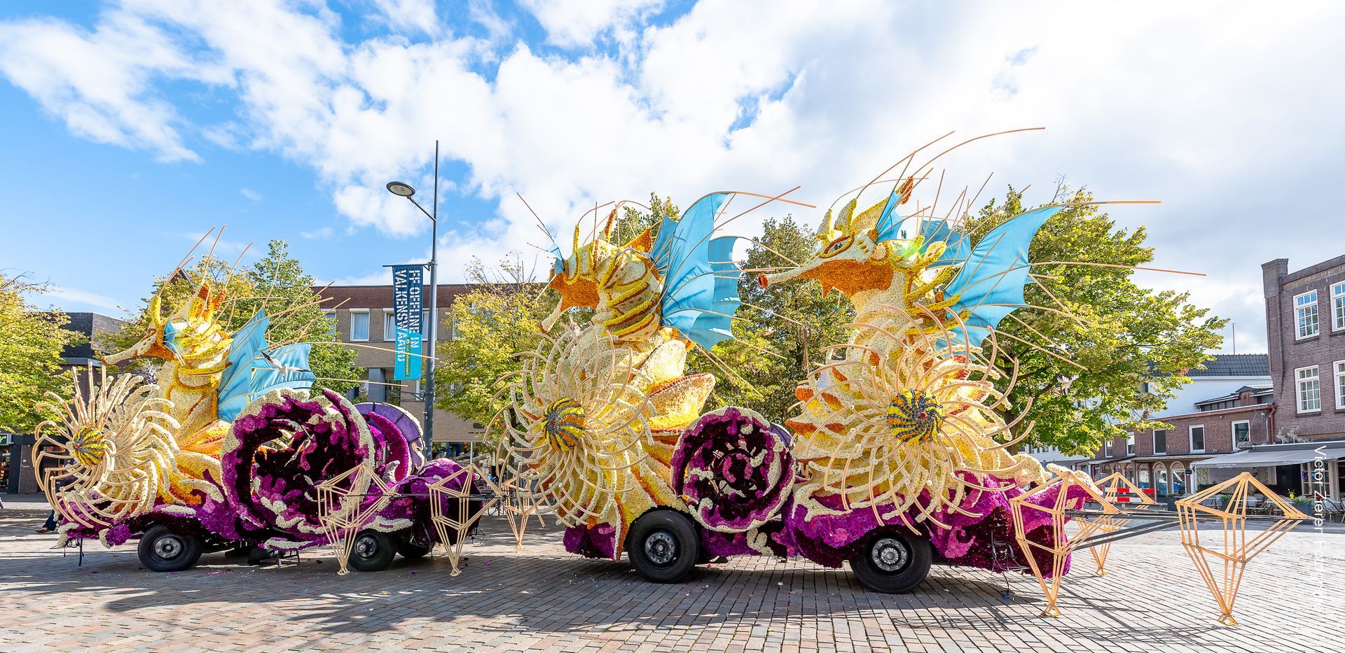 Kleurrijke praalwagens, versierd met bloemen en veren, rijden door een stadsstraat onder een blauwe hemel.