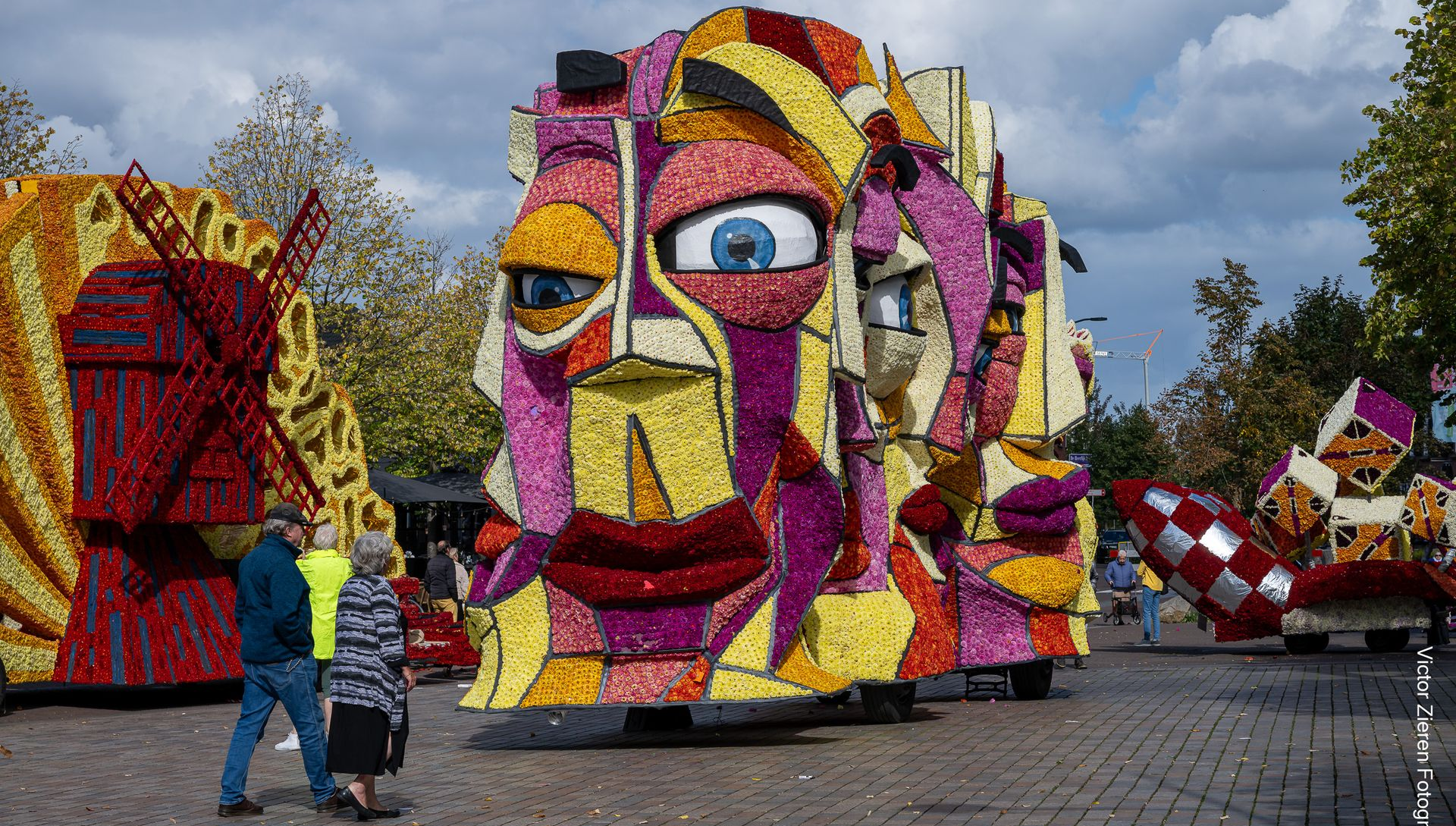 Mensen staan naast een kleurrijk mozaïekgezicht dat buiten onder een bewolkte hemel tentoongesteld wordt.