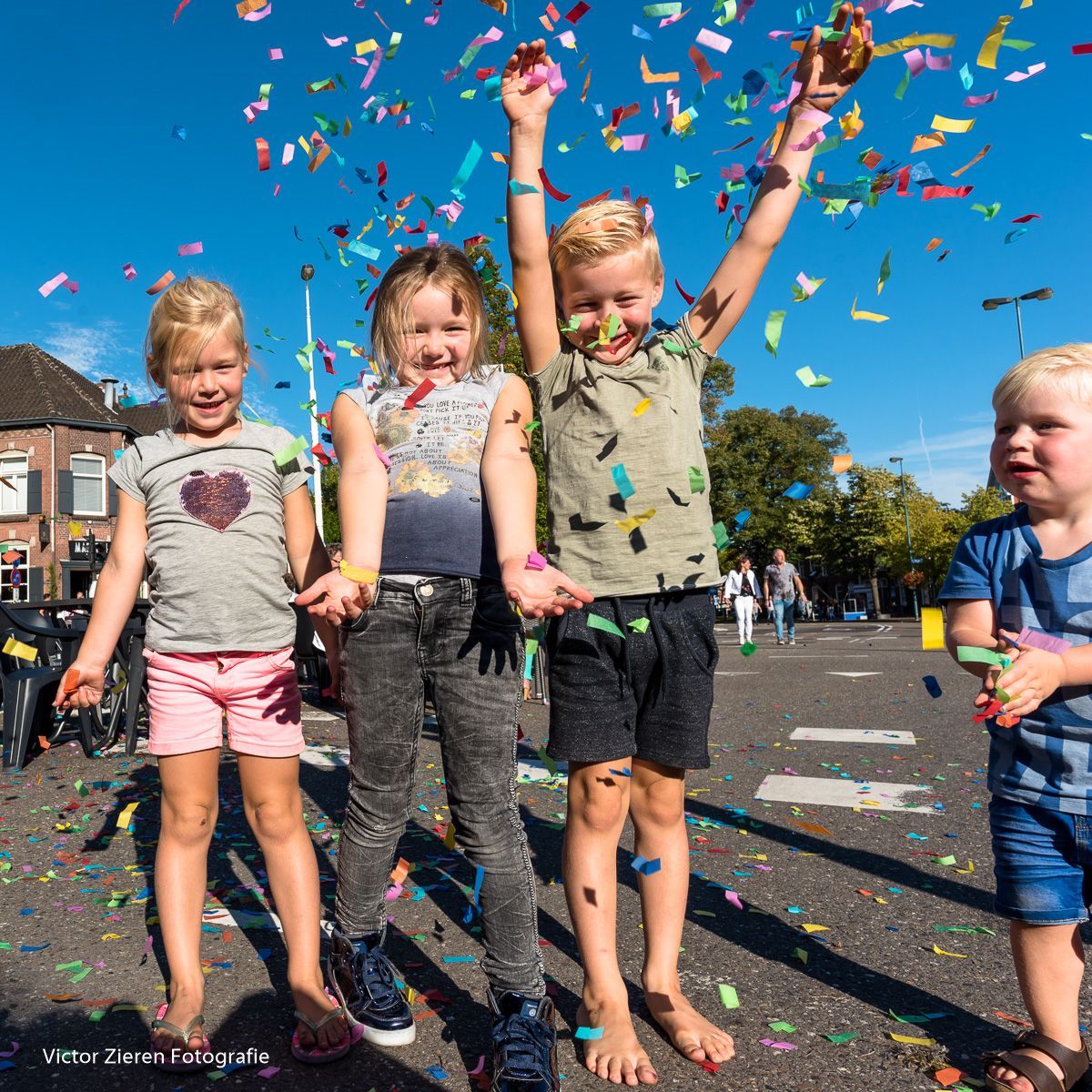 Kinderen onder kleurrijke confetti op een zonnige straat, lachend en hun armen in de lucht stekend om feest te vieren.