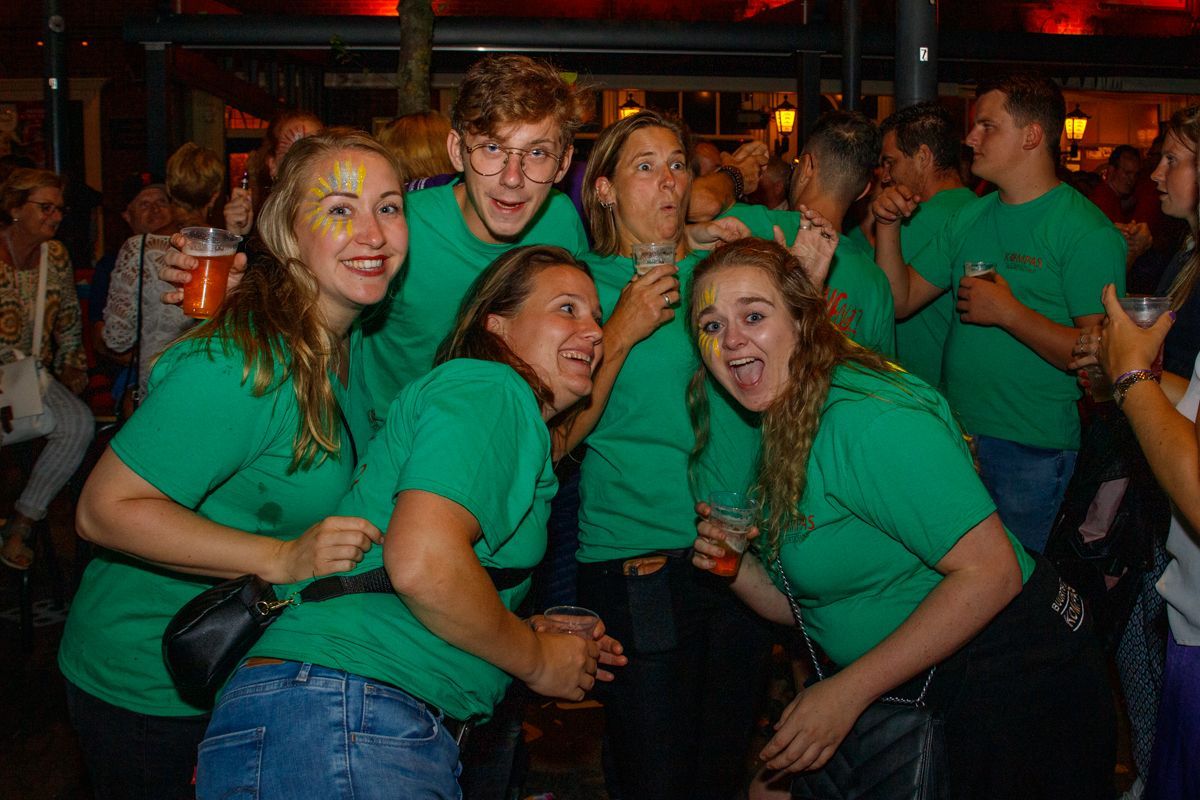 Een groep mensen in groene shirts poseert en lacht tijdens een evenement in een schemerig verlichte binnenruimte.