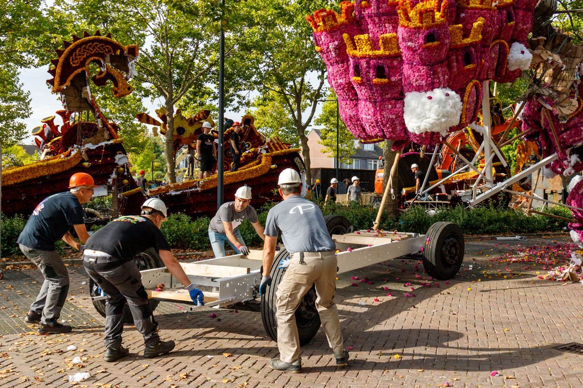 Arbeiders verplaatsen een grote, met bloemen versierde praalwagen met felroze wielen in een park.