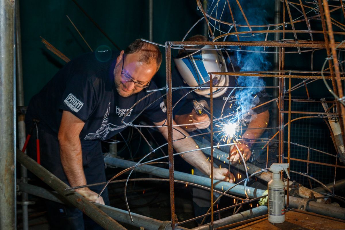 Arbeiders lassen metalen steigers in een schemerige industriële omgeving, met felle vonken en blauw licht.