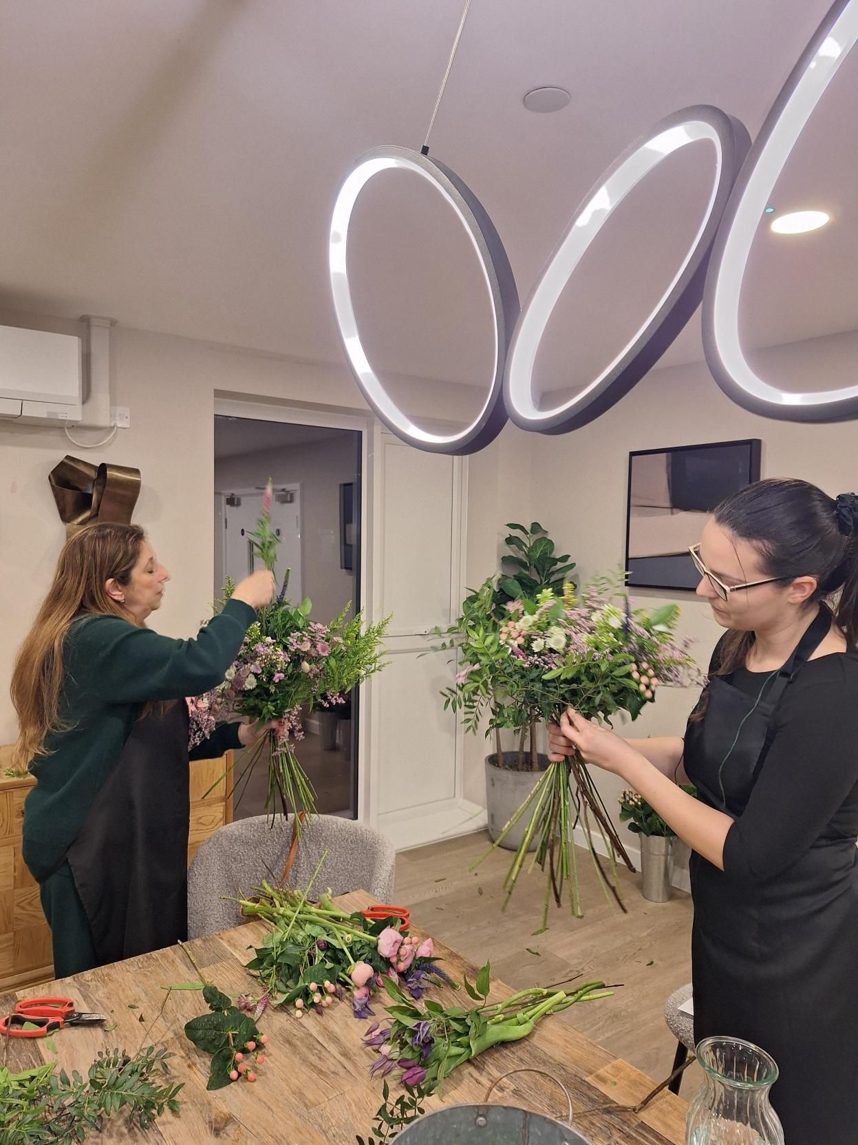 Two people arranging flowers around a wooden table under glowing oval ceiling lights in a workshop room