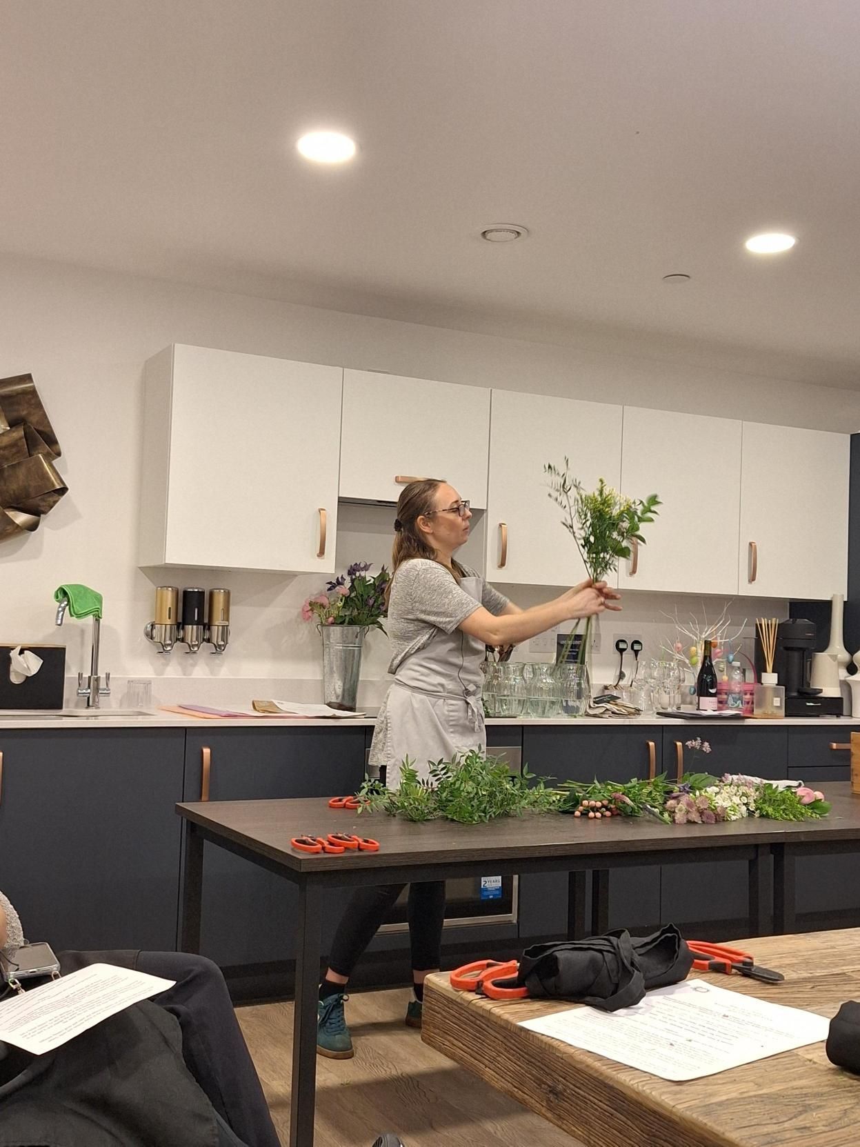 Person arranging flowers in a bright kitchen with tables, plants, and supplies.