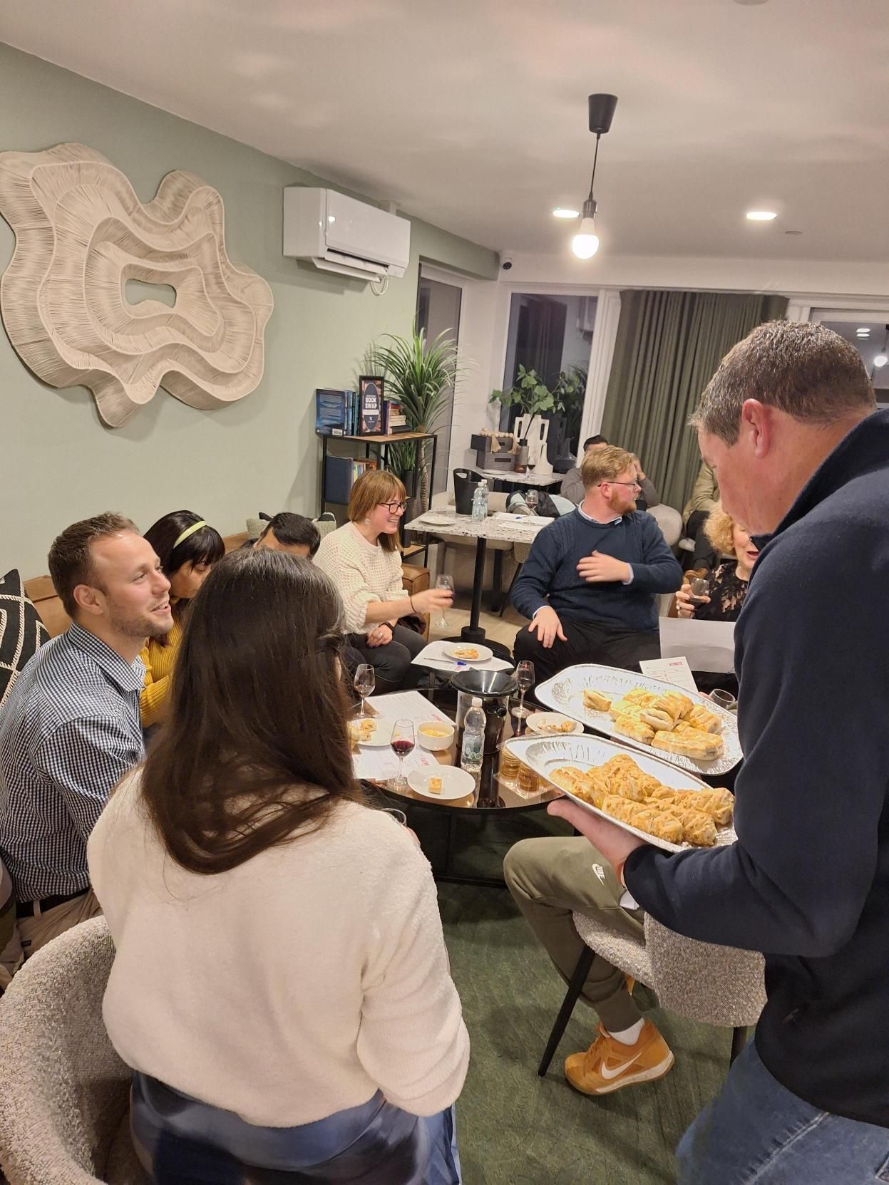 People gathered around a table at a home gathering, serving and eating food in a bright kitchen.