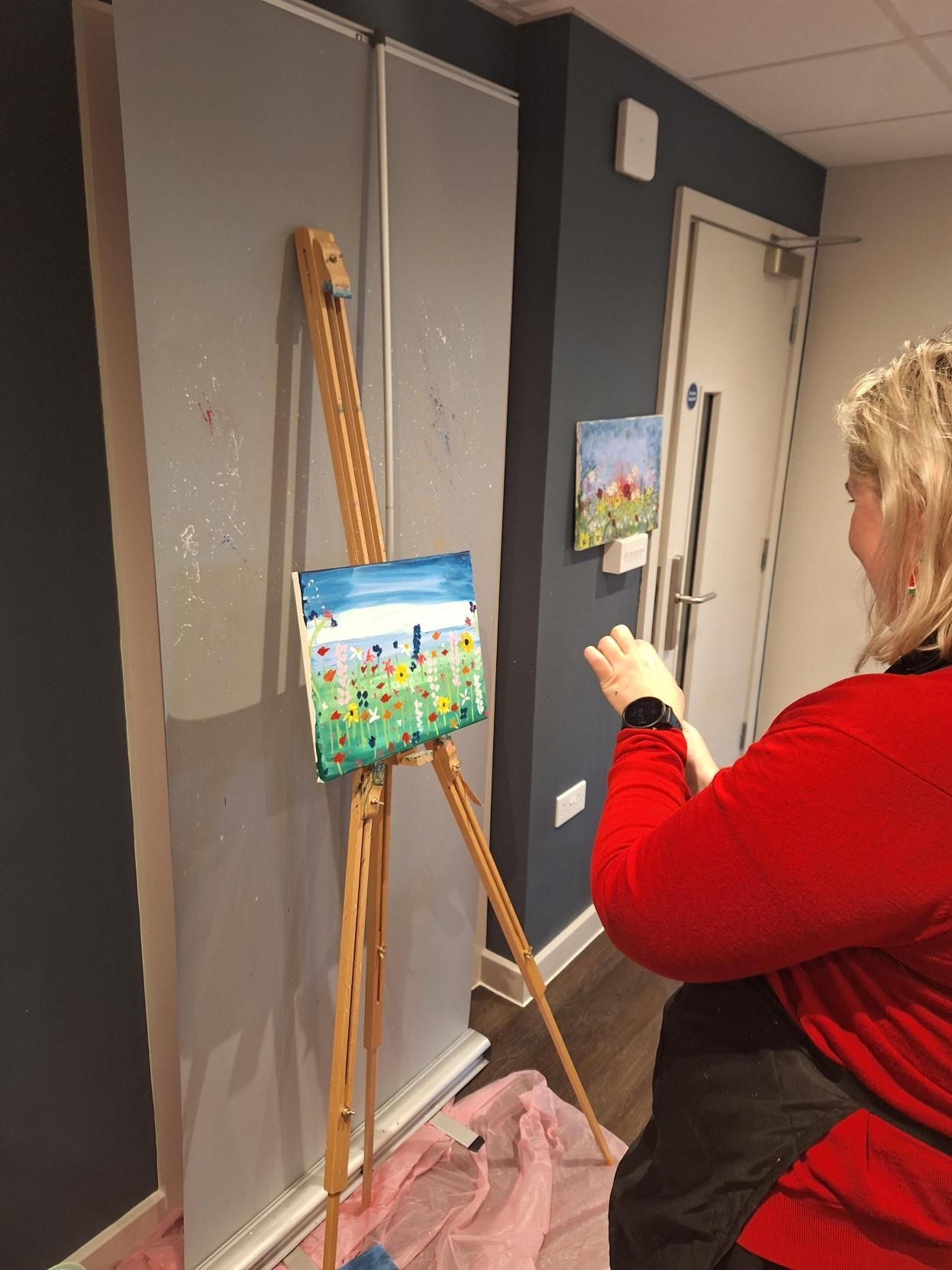 Person painting a small landscape on an easel in a hallway, with a ladder leaning nearby.