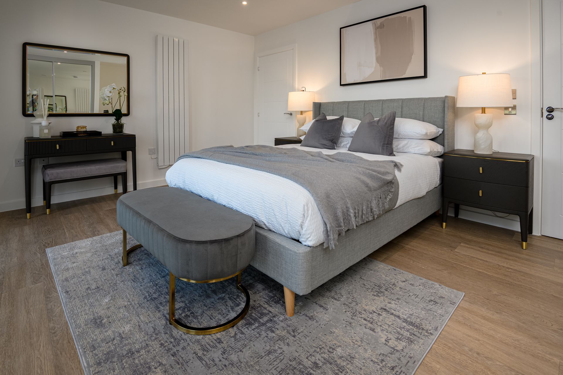 Modern bedroom with gray bed, matching ottoman, black nightstands, vanity, and light wood floors