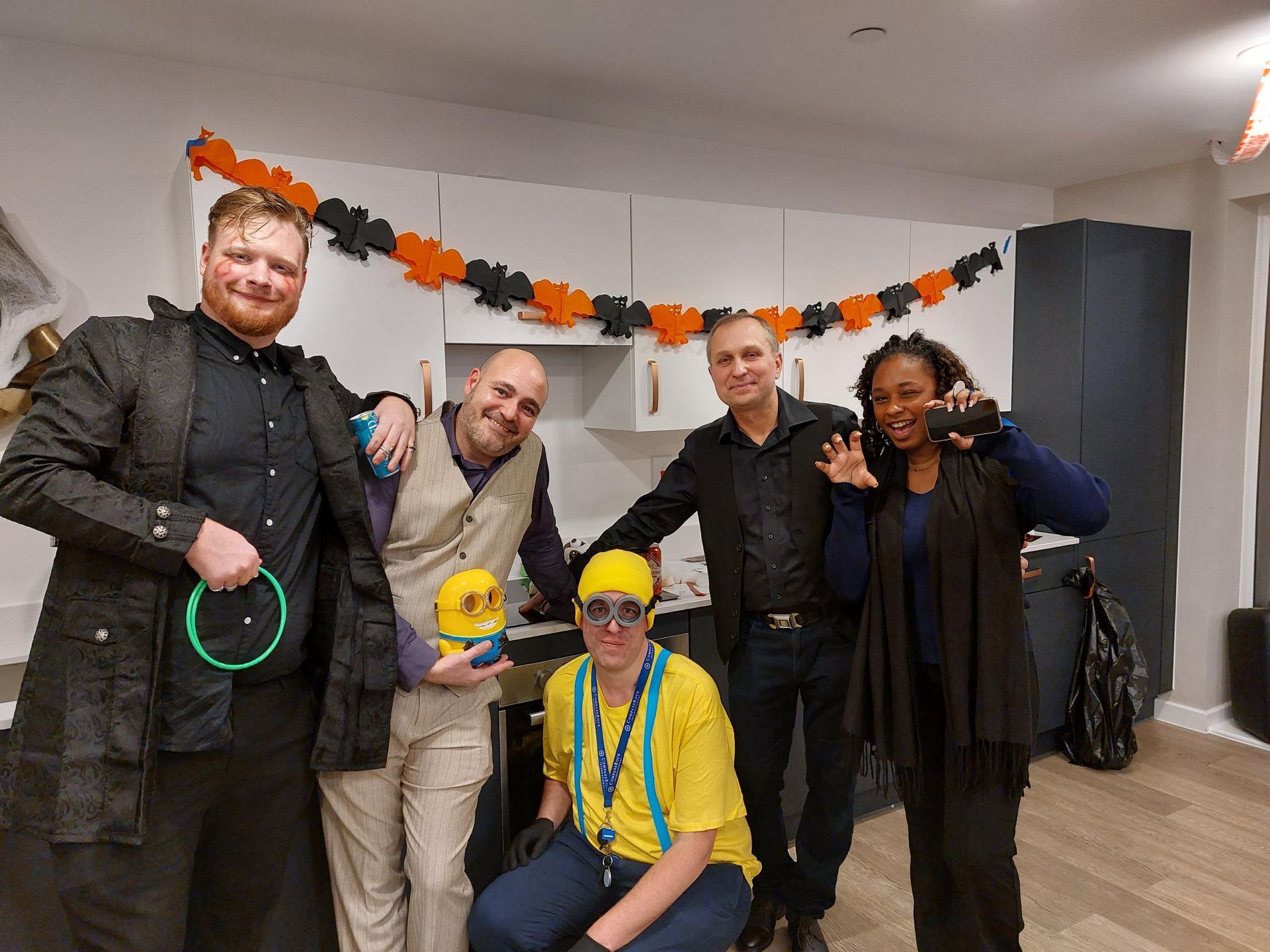 Five people posing indoors at a Halloween party, with fall garland and costumes.