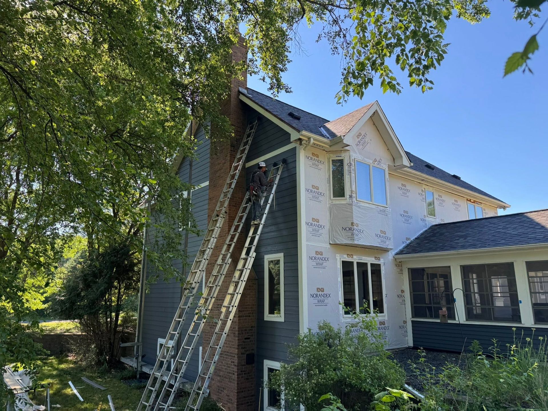 Ladder leaning against a two-story house with gray siding and white trim under leafy trees