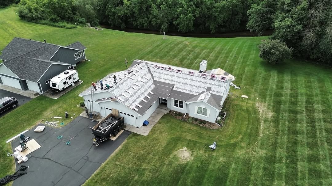Aerial view of a house under construction beside a paved driveway and green lawn, with construction equipment nearby