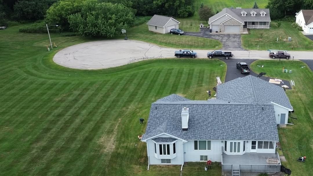 Aerial view of a house with a striped lawn, driveway, and a baseball field beyond the yard