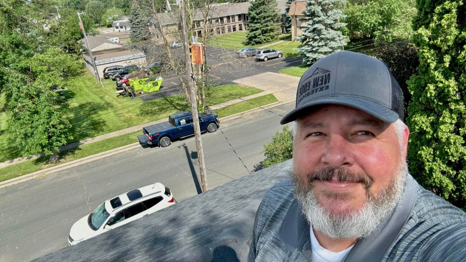 Smiling man in gray cap on a rooftop, overlooking a street with parked cars and trees below
