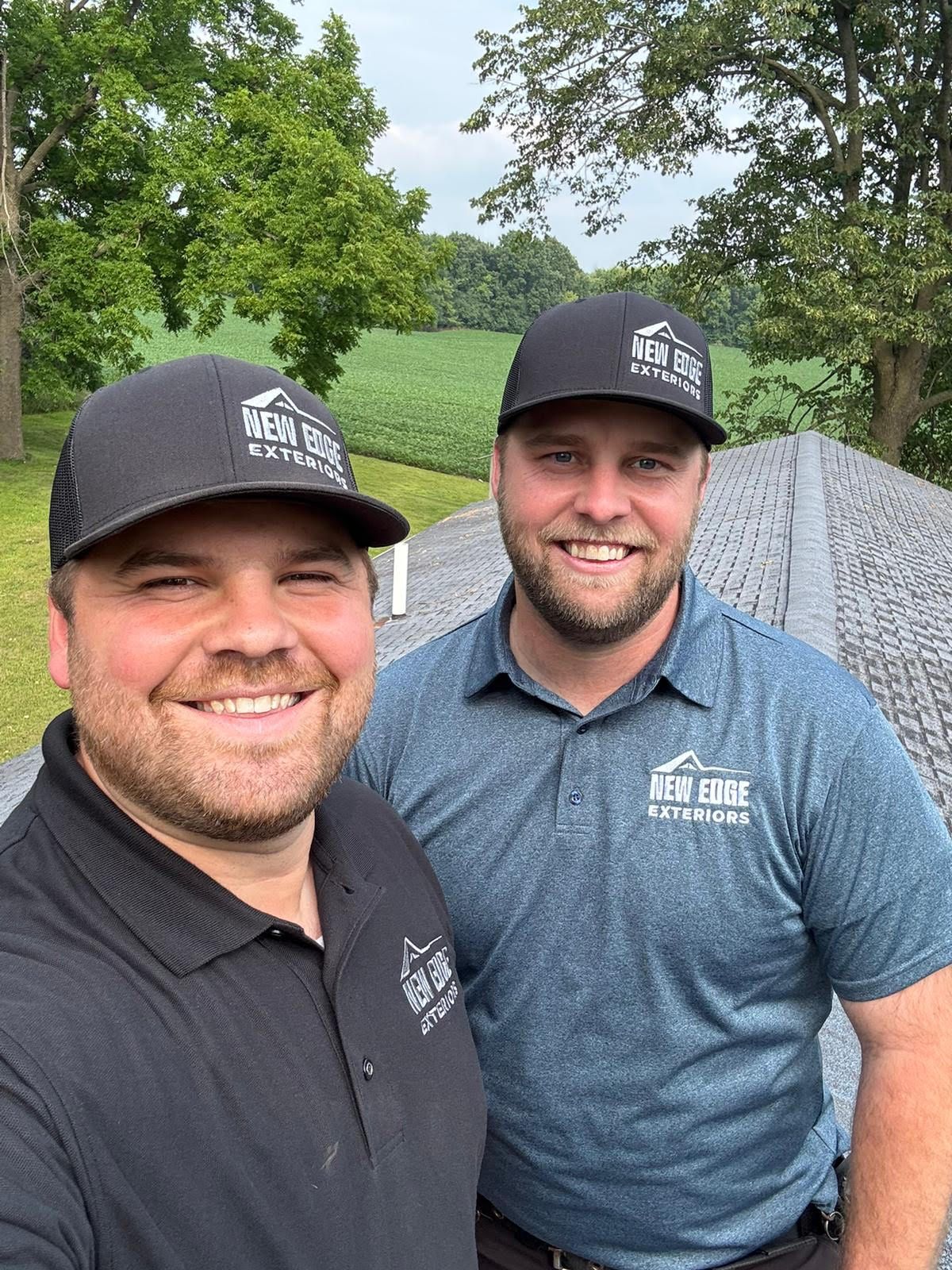Two men in black and blue polos smiling outdoors by a grassy field under trees