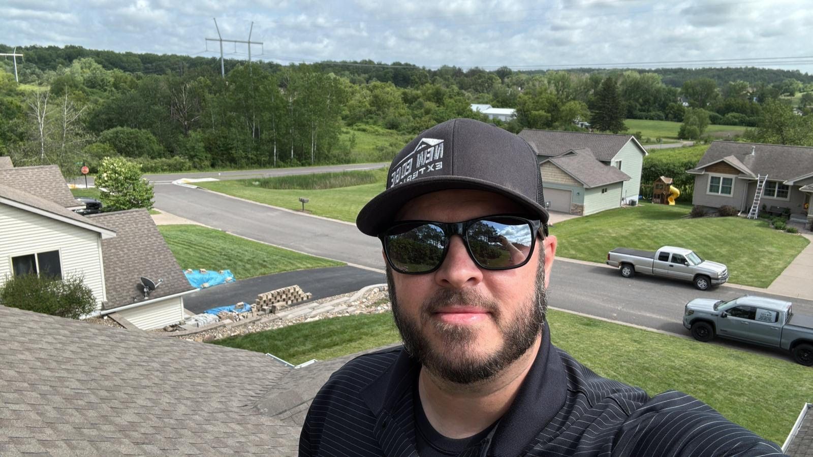Man wearing sunglasses and a cap takes a selfie on a rooftop, with suburban houses and trees in the background.