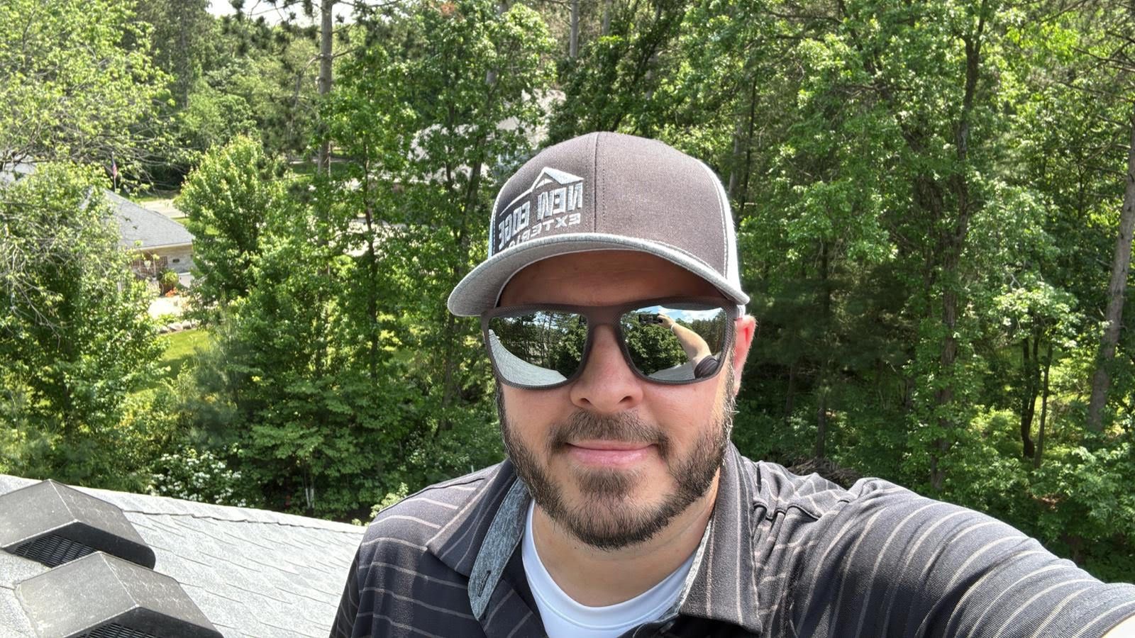 Selfie of a person wearing a cap and sunglasses on a rooftop with trees in the background