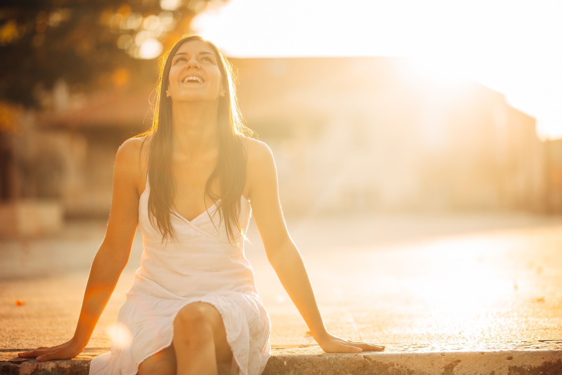 A woman in a white dress is sitting on the sidewalk looking up at the sun.