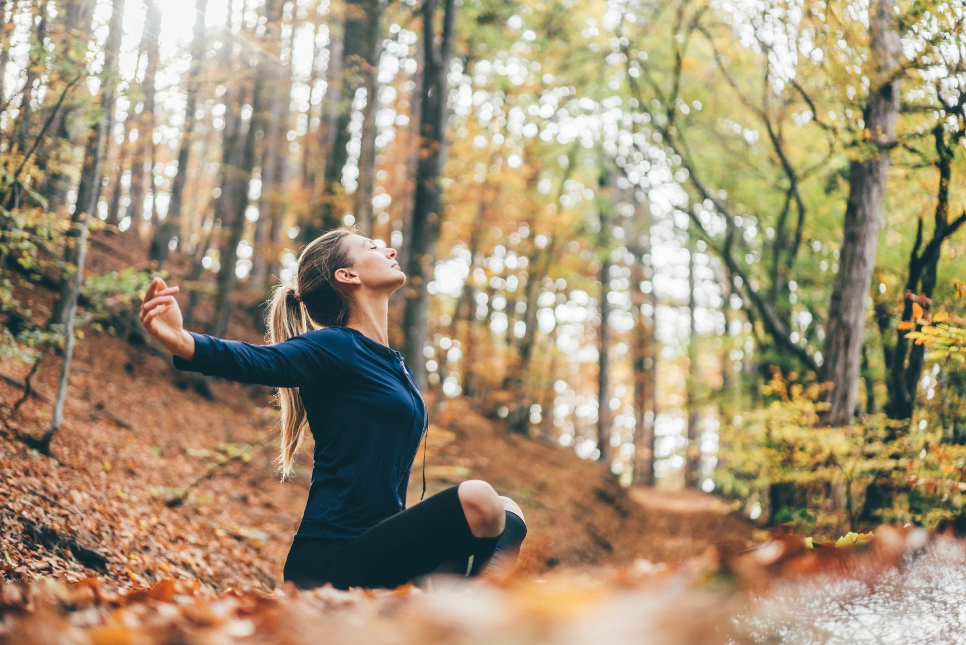 A woman is sitting on the ground in the woods with her arms outstretched.