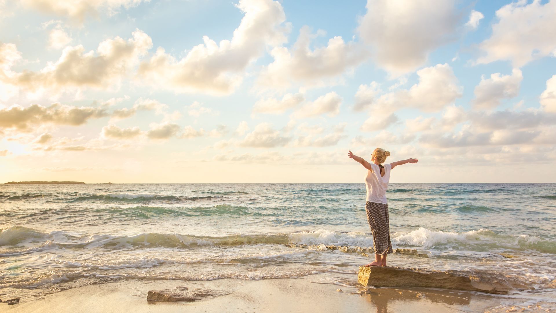 A woman is standing on a rock on the beach with her arms outstretched.