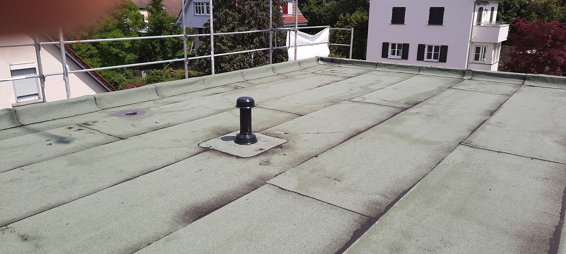 Flaches graues Dach mit kleinem Entlüftungsrohr, mit Blick auf Bäume und Wohnhäuser