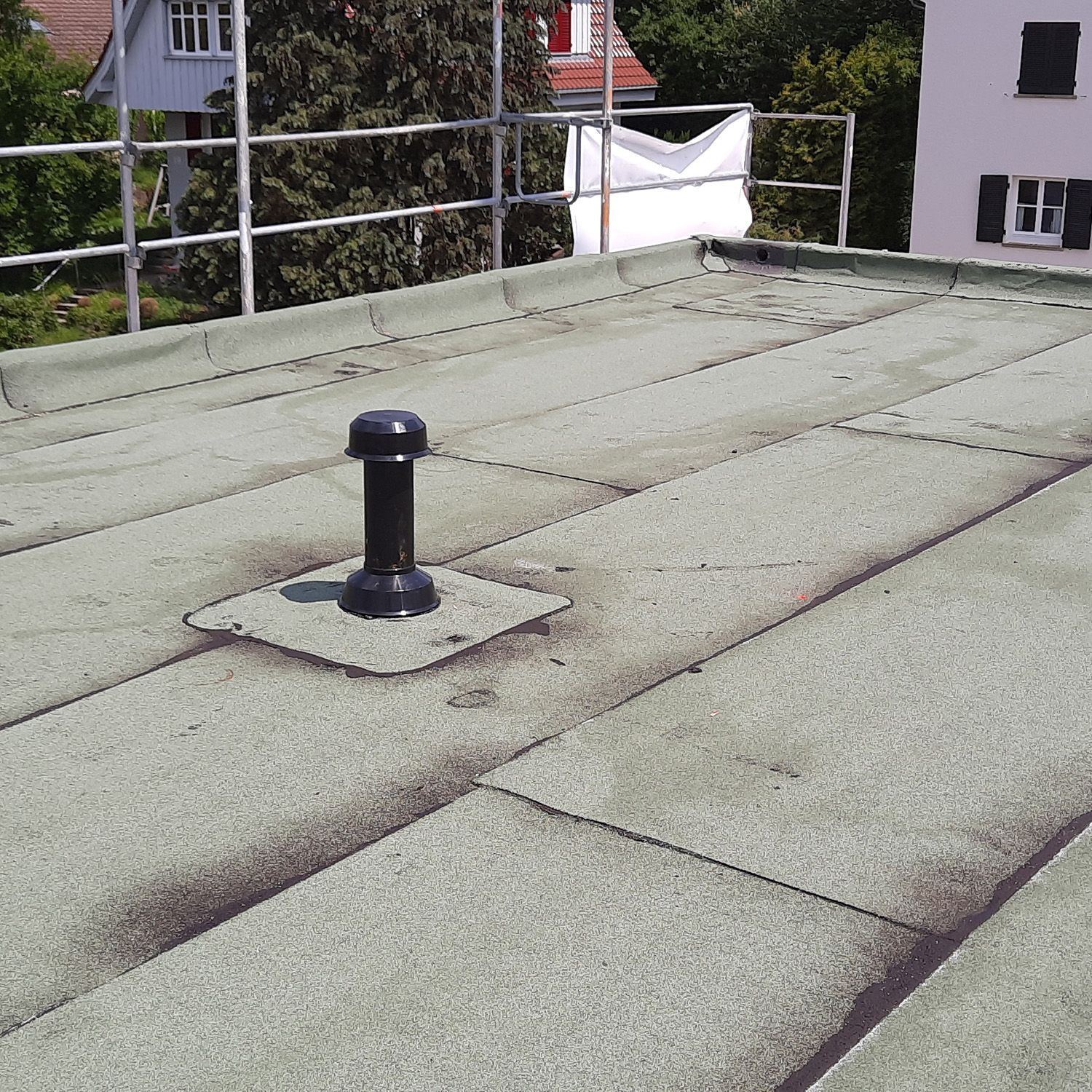 Flachdach mit schwarzem Entlüftungsrohr, Kiesoberfläche und Sicherheitsgeländer am Rand.