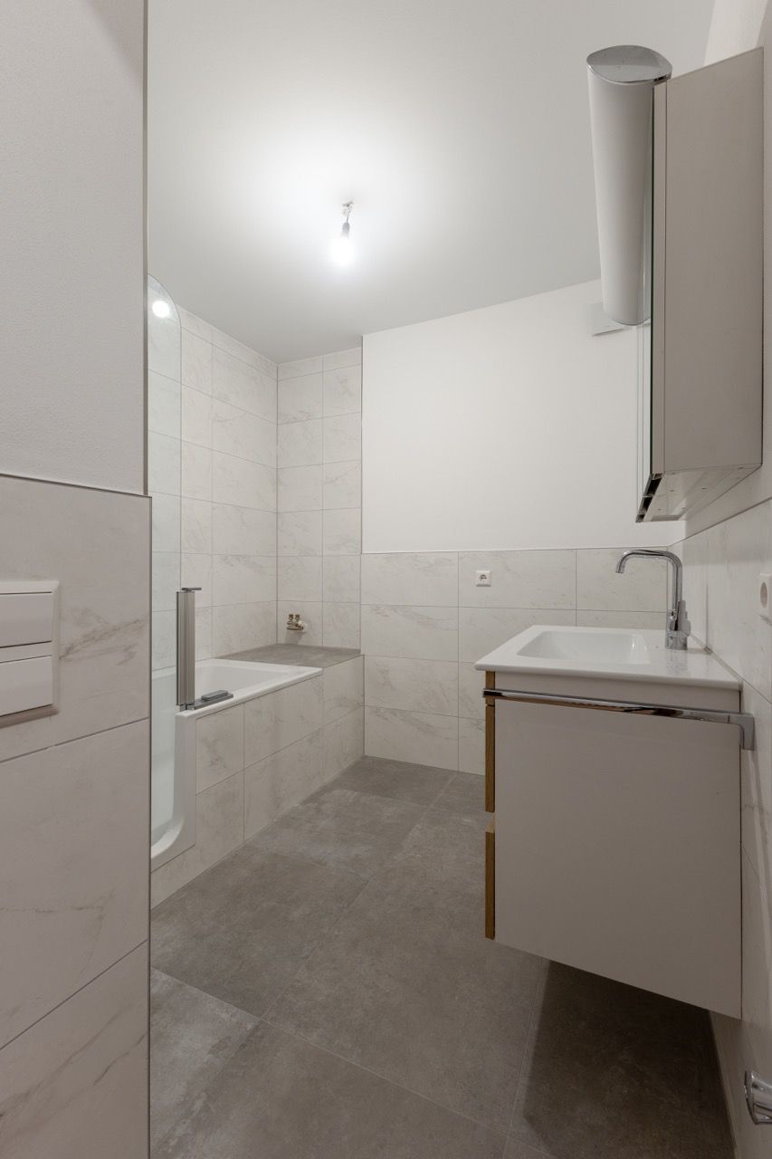Kleines weißes Badezimmer mit Badewanne, Waschtischunterschrank, Spiegel sowie gefliesten Wänden und Boden.